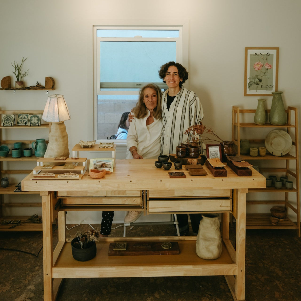 Two women standing behind a wooden table with various items in a room with shelves and decor.